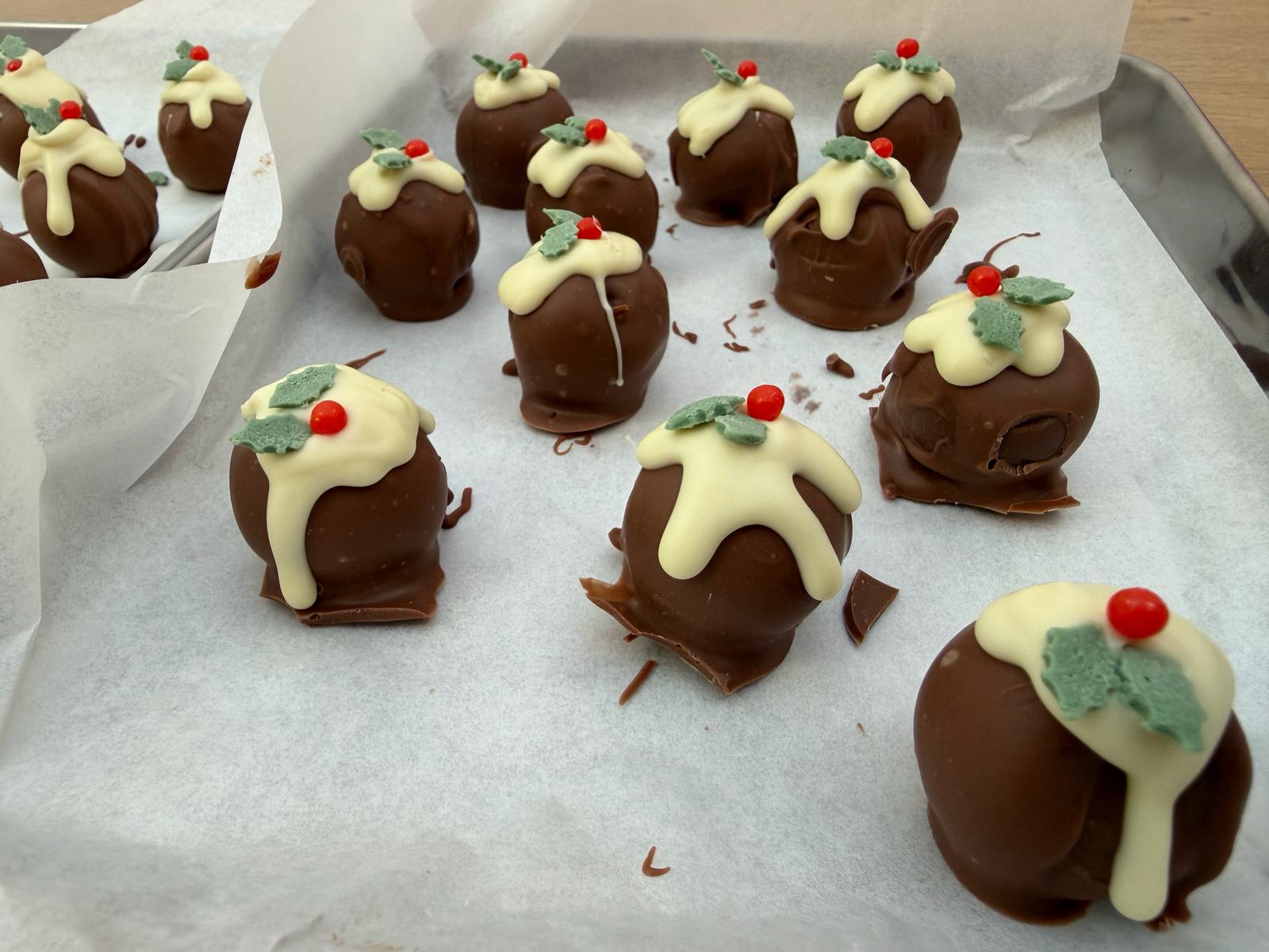 A tray of handmade Christmas pudding truffles: round chocolate truffles with white chocolate icing dripping down the sides, topped with green holly leaves and red berries